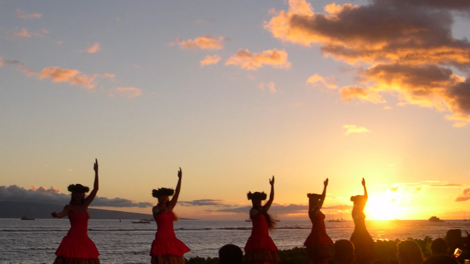 Hula dancers in red dresses raise their arms in silhouette against a blazing orange sunset over the ocean, with luau guests seated at tables in the foreground