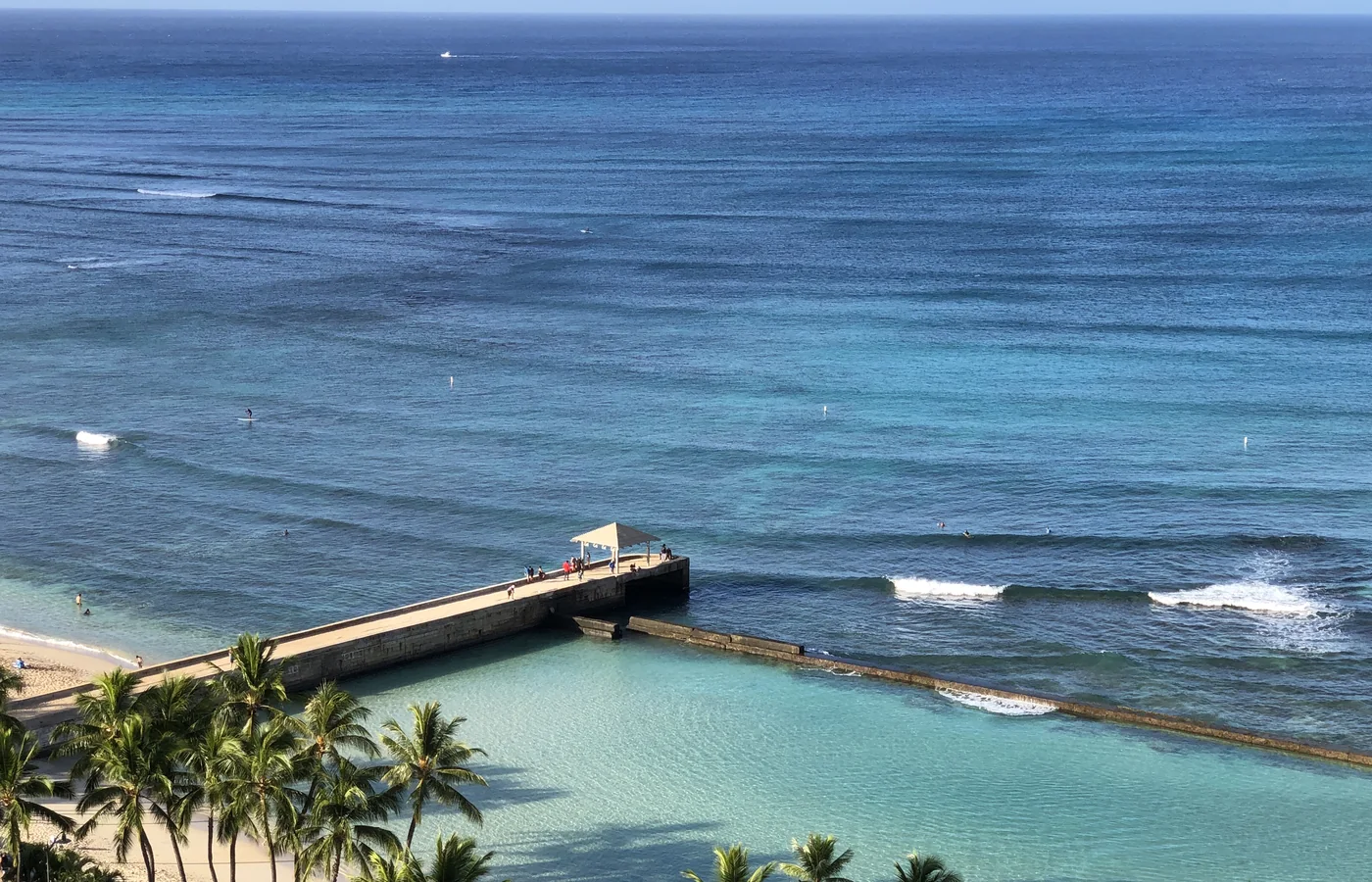 A turquoise lagoon and the open Pacific seen from a high balcony above Waikiki, with a stone pier, surfers and palm trees in the foreground